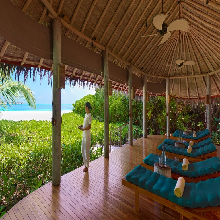 Wooden deck with lounge chairs beneath a thatched roof, overlooking green landscape and ocean, designed for relaxation and connection to nature.