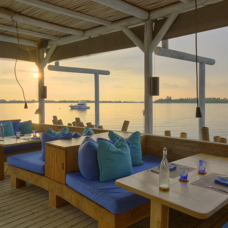 Restaurant terrace with wooden tables and blue cushioned chairs, facing tranquil sea views, distant boats, and an island on the horizon at dusk.