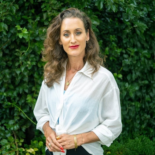 Individual with wavy brown hair, a white button-up shirt, and gold jewelry stands near vibrant green plants, meeting the viewer’s gaze.