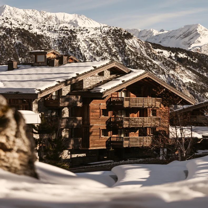 Chalet featuring balconies among snow-covered rooftops, with pine forests and mountains in the distance beneath a clear sky.
