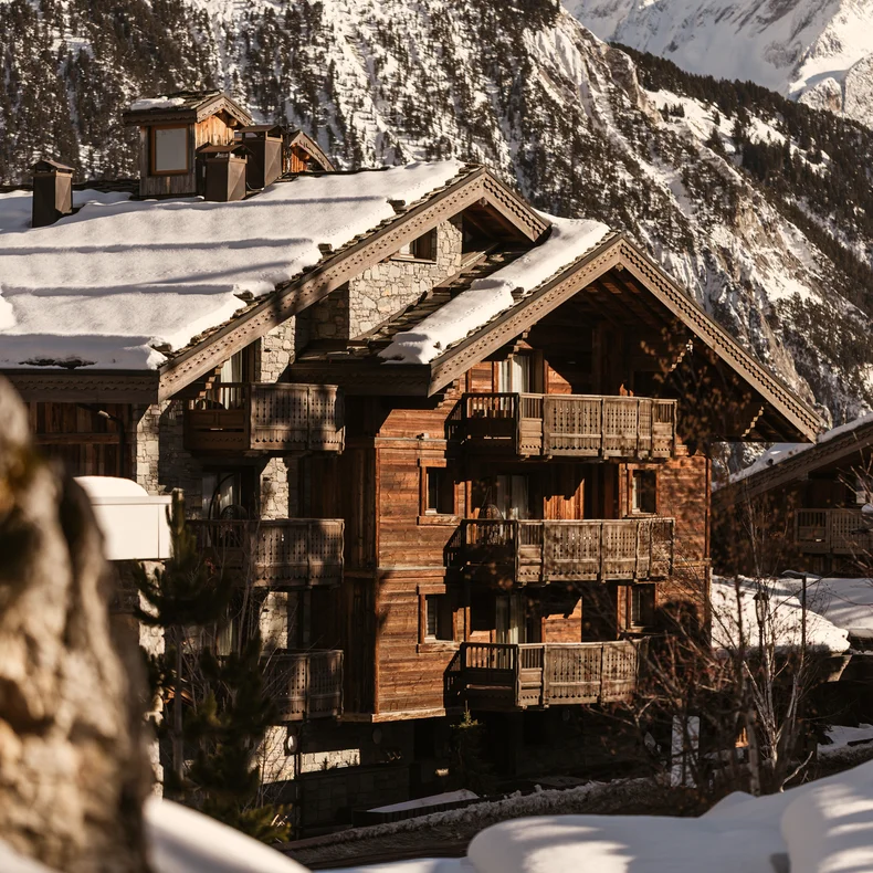 Chalet featuring balconies among snow-covered rooftops, with pine forests and mountains in the distance beneath a clear sky.