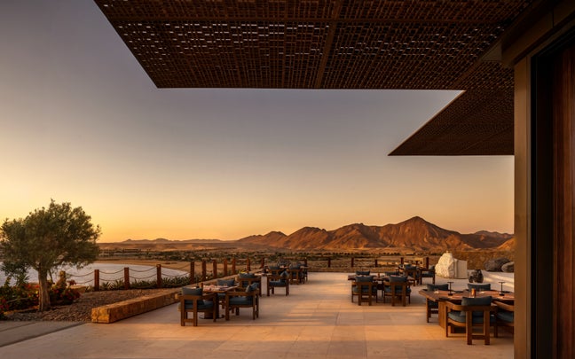 Restaurant terrace with orderly tables and chairs, offering views of mountains and water under a clear sky. Accessible pathways ensure comfortable navigation.