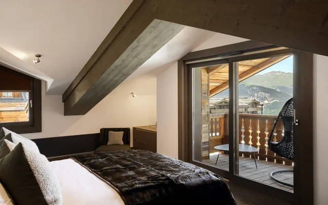 Chambre à coucher sereine avec des matériaux durables, des textiles chauds, un plafond en bois et des portes vitrées menant à un balcon avec vue sur les montagnes et des sièges extérieurs.
