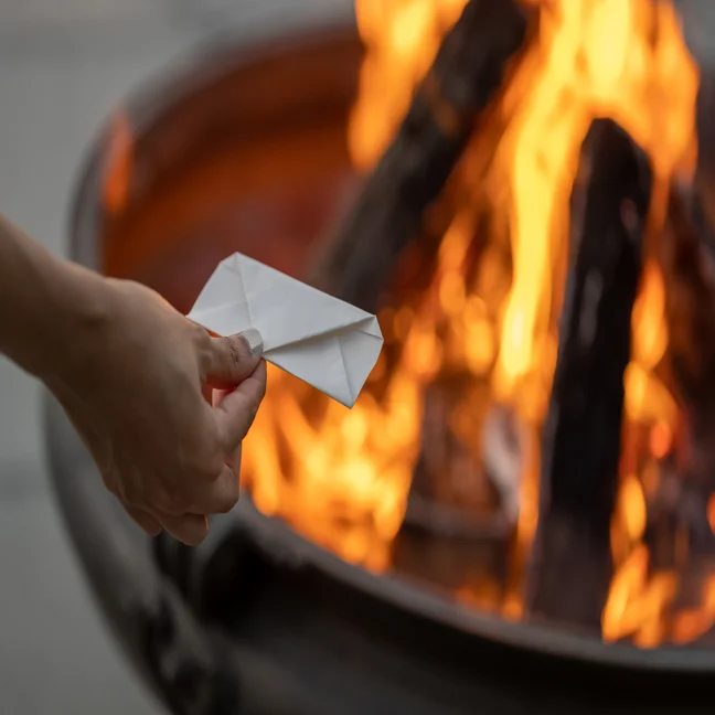 A person prepares to place a folded paper envelope into the flames of an outdoor fire pit, highlighting mindful actions and a connection to natural elements.