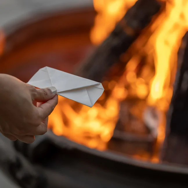 A person prepares to place a folded paper envelope into the flames of an outdoor fire pit, highlighting mindful actions and a connection to natural elements.