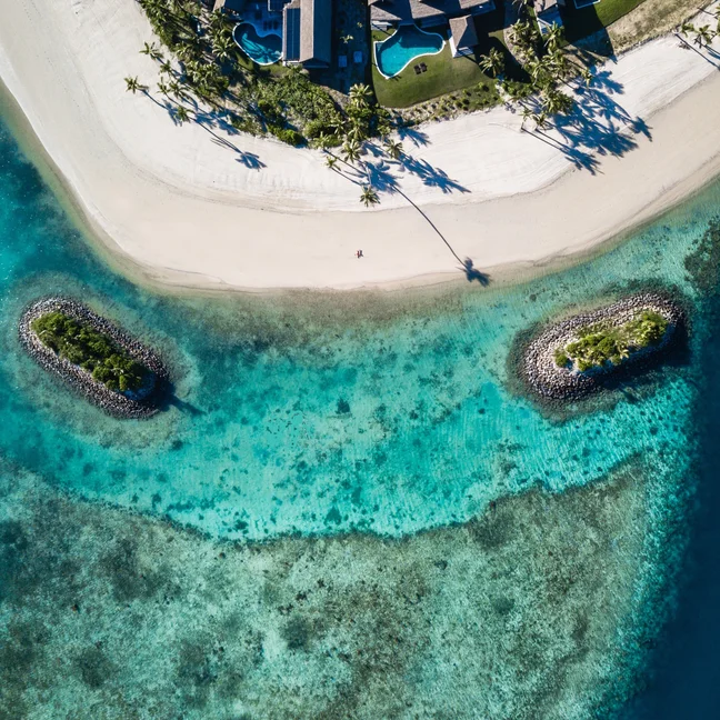 Beachside villas featuring private pools, soft white sands, clear blue water, and two lush green islands just off the tranquil shoreline.