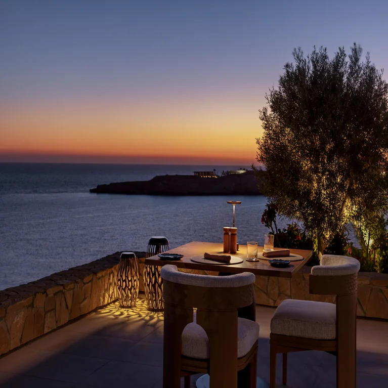 Outdoor dining area with a lit tree, stone wall, modern chairs, and sea view at dusk under a tranquil sky. Accessible pathways and clear seating arrangement.
