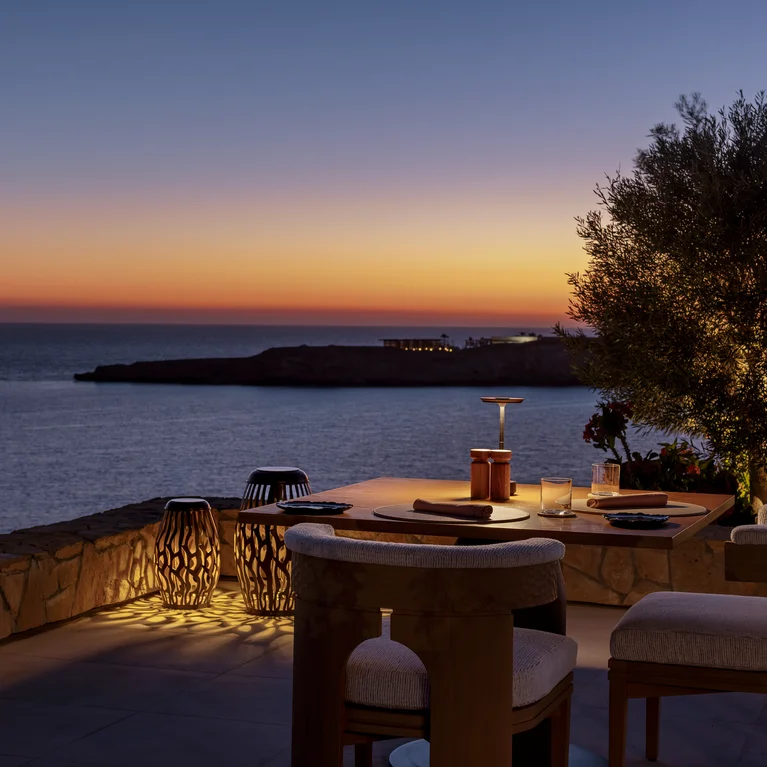 Outdoor dining area with a lit tree, stone wall, modern chairs, and sea view at dusk under a tranquil sky. Accessible pathways and clear seating arrangement.