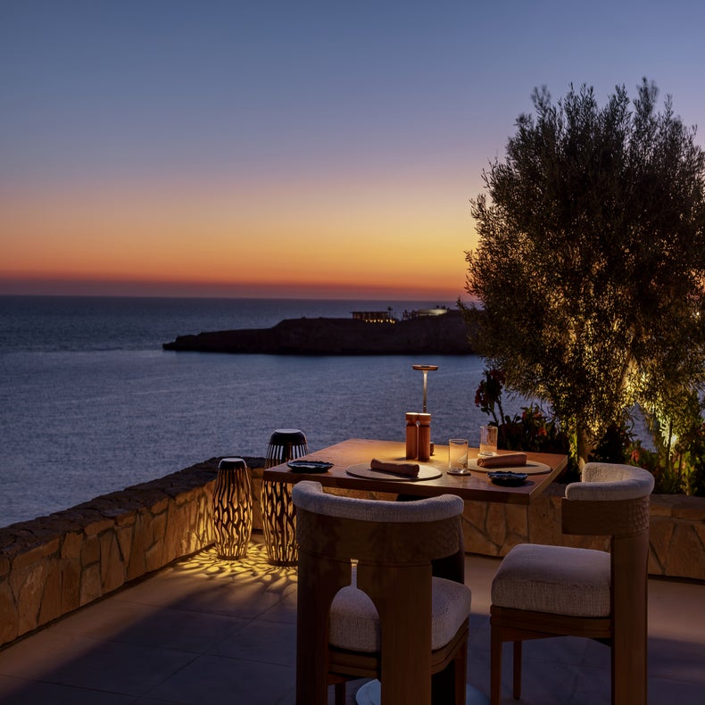 Outdoor dining area with a lit tree, stone wall, modern chairs, and sea view at dusk under a tranquil sky. Accessible pathways and clear seating arrangement.