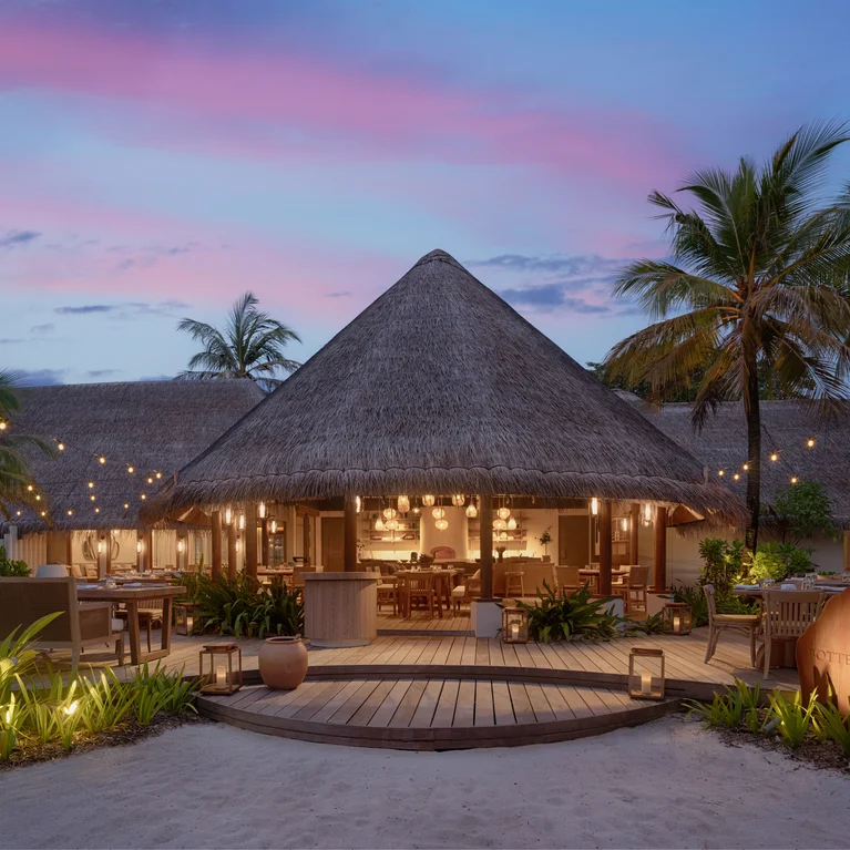 غرفة طعام مفتوحة في الهواء الطلق مضاءة بأضواء خيطية، مع مقاعد خشبية على التراس، محاطة بأشجار النخيل والنباتات الاستوائية عند غروب الشمس.