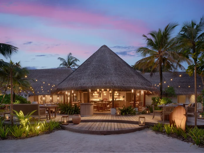 Open-air dining space illuminated by string lights, with wood deck seating among palm trees and tropical vegetation at dusk.