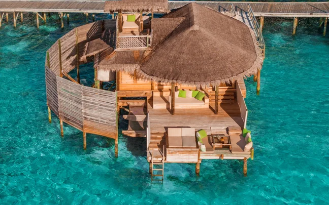 Wooden villa with thatched roof, private deck and accessible boardwalk, set above clear ocean waters with space for relaxation and connection to the outdoors.