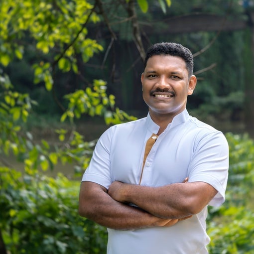 Person in a white shirt stands outdoors, arms crossed and smiling, surrounded by greenery and trees with a softly blurred bridge behind.
