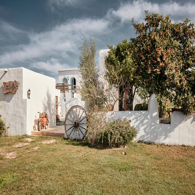 A white Mediterranean-style building features a wagon wheel, surrounded by greenery and trees beneath a partly cloudy sky.
