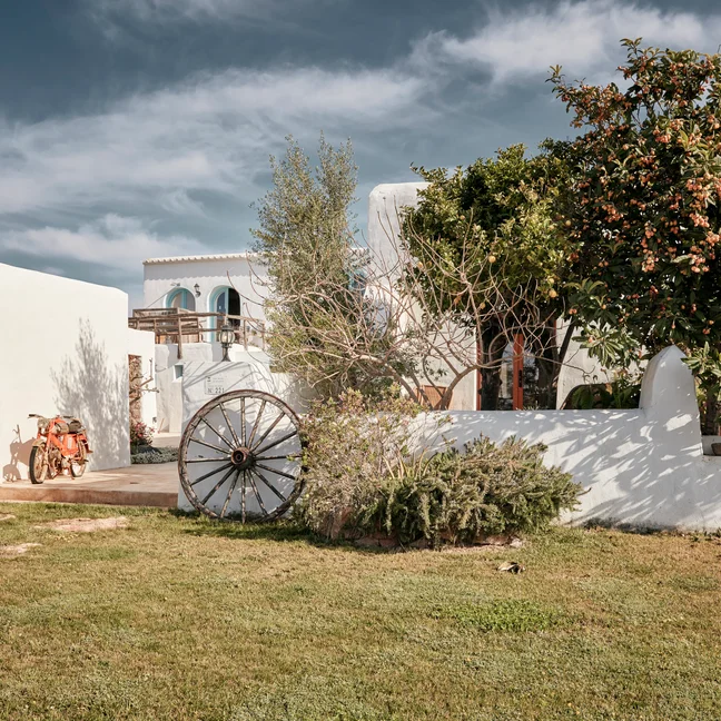A white Mediterranean-style building features a wagon wheel, surrounded by greenery and trees beneath a partly cloudy sky.