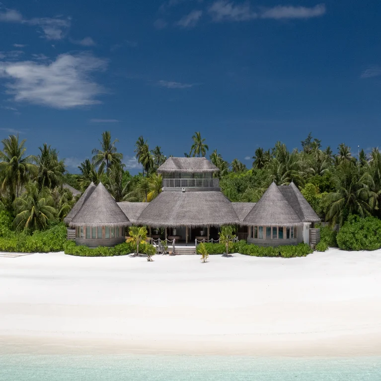 Beachfront villa with thatched roofs among palm trees and native plants, overlooking white sand and blue-green sea under an open sky.