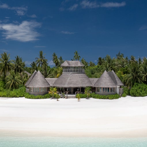 Beachfront villa with thatched roofs among palm trees and native plants, overlooking white sand and blue-green sea under an open sky.