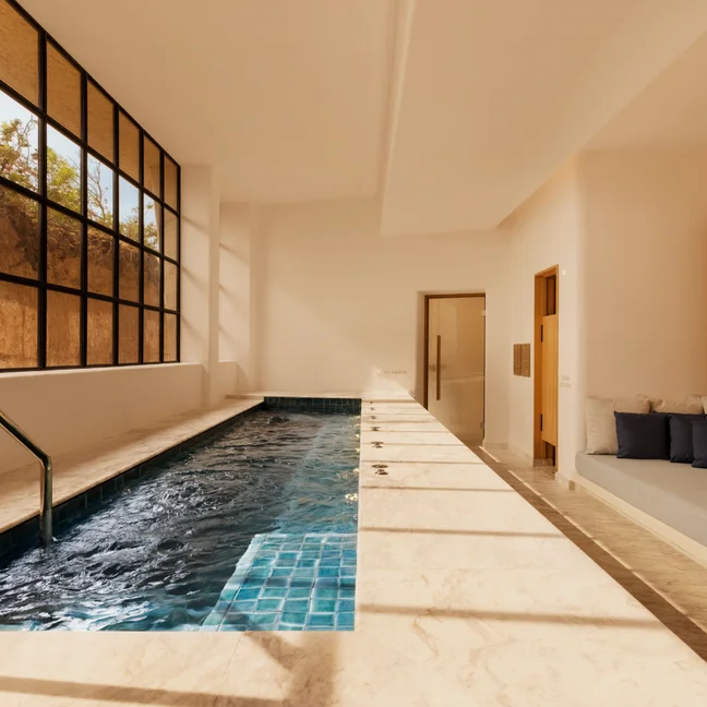 Indoor pool featuring blue tiles, expansive windows for abundant natural light, marble flooring, and cushioned seating along one side. Clean design supports relaxation.