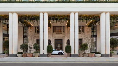 Building entrance with tall columns, abundant greenery above the doorway, potted plants, statues by the entrance, and a car parked at the front.