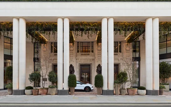 Building entrance with tall columns, abundant greenery above the doorway, potted plants, statues by the entrance, and a car parked at the front.