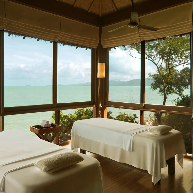 غرفة سبا مع طاولتين للتدليك، ونوافذ كبيرة تطل على البحر والنباتات المحيطة بها، مصممة للعافية والاسترخاء الواعي.