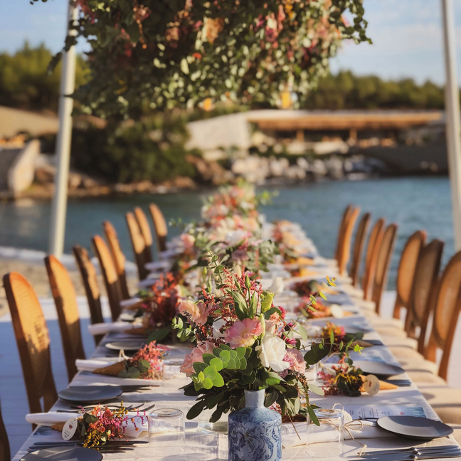Outdoor table arranged with fresh floral centerpieces, dining settings, and wooden chairs; peaceful water and lush plants form the natural backdrop.