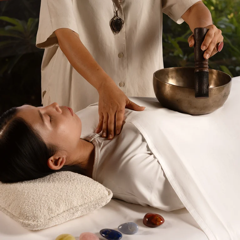 يتلقى أحد الضيوف جلسة علاج صوتي، وهو مستلقٍ على طاولة تدليك مغطاة بملاءة بيضاء، بينما يحمل شخص آخر وعاء غناء تبتي فوق رأسه. توضع أحجار الشفاء في مكان قريب.