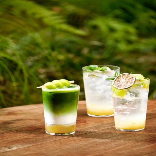 Tall glasses with vibrant, layered drinks topped with lime, herbs, and melon balls on a wooden table outside, surrounded by lush green foliage.