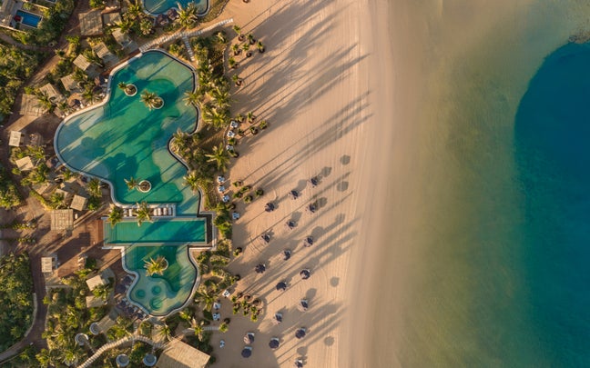 Resort pool bordered by palm trees, sandy beach, and calm blue ocean; lounge chairs and umbrellas spaced for relaxation amid natural surroundings.