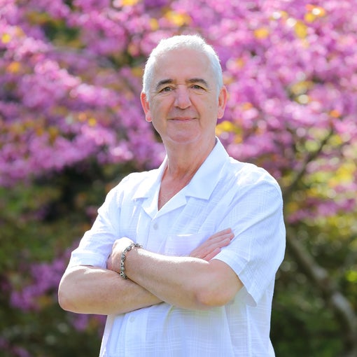 Man with short gray hair stands outside, arms gently crossed, smiling in a white shirt. A pink flowering tree and lush greenery are softly blurred in the background.