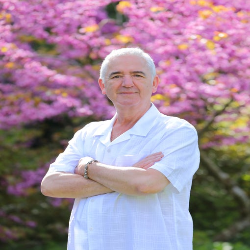 Man with short gray hair stands outside, arms gently crossed, smiling in a white shirt. A pink flowering tree and lush greenery are softly blurred in the background.