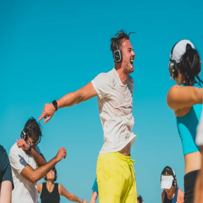 Guests with headphones enjoy outdoor movement beneath a blue sky; a person in yellow shorts stands at the center, arms open and smiling.