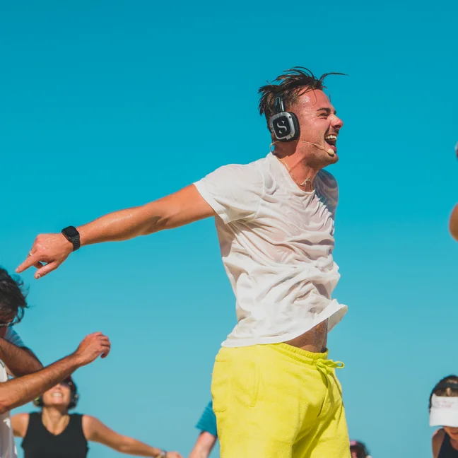 Guests with headphones enjoy outdoor movement beneath a blue sky; a person in yellow shorts stands at the center, arms open and smiling.
