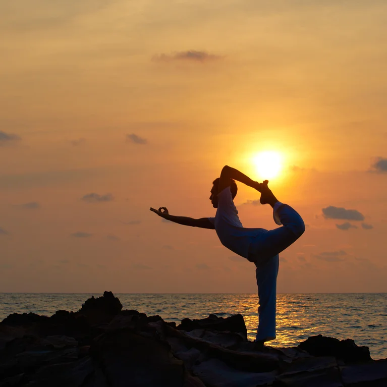 Individual holding a yoga pose on seaside rocks at dusk, framed by an amber sky and low sun, embodying balance and connection to nature.