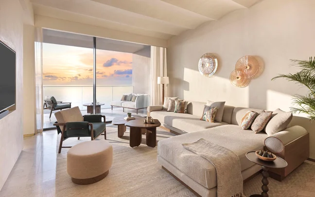 Serene living area with soft-toned furnishings, generous seating, curated artwork, and doors leading to a balcony with an ocean view at dusk.