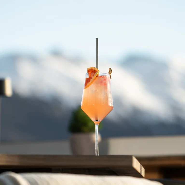 户外餐桌上摆放着一杯粉红色的鸡尾酒,配上一片橘子和一根吸管,背景是白雪皑皑的群山,稍稍有些模糊。.