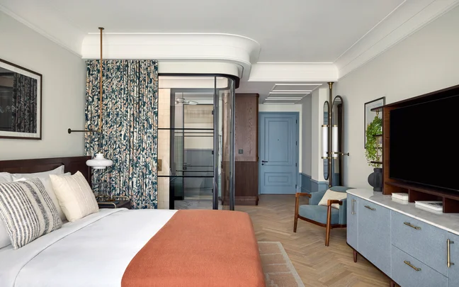 Serene room featuring a bed with natural textiles, soothing blue armchair, custom wood floors, accessible glass shower, and calming accents in blue throughout.