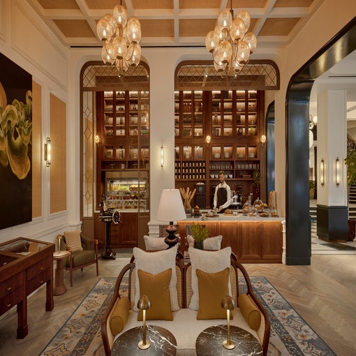 Lounge with soft seating, ambient lighting, glass light fixtures, wooden shelves of beverages, and a team member preparing fresh food at a counter.