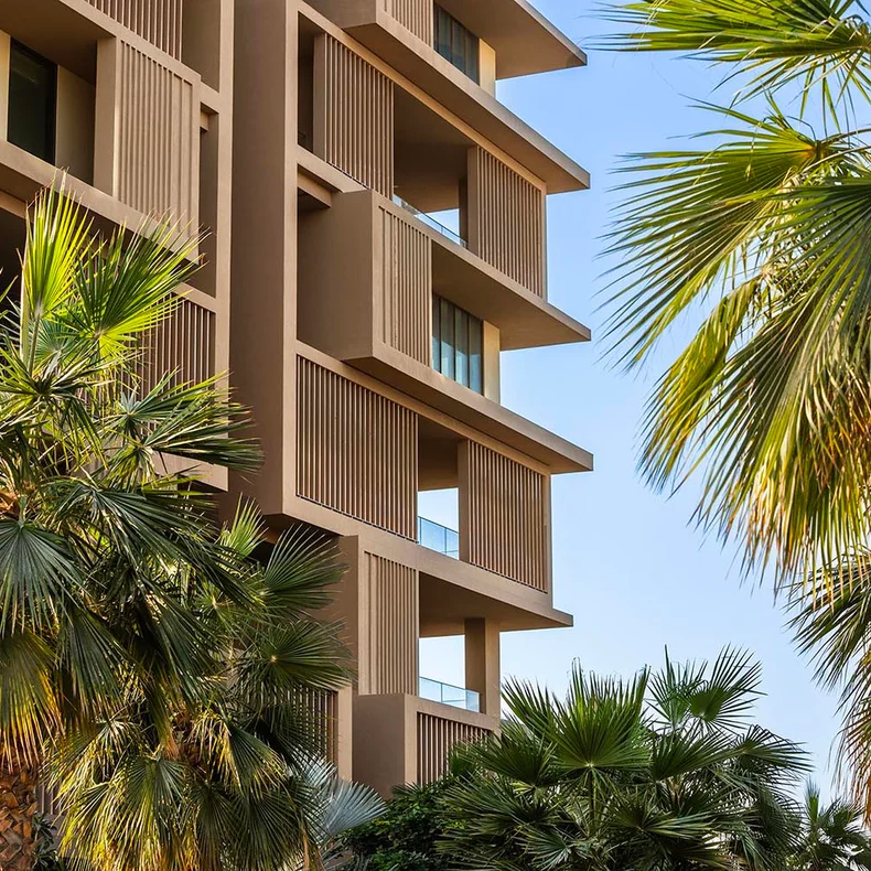Contemporary building featuring spacious outdoor terraces and vertical wooden elements, framed by palm trees against a bright sky.