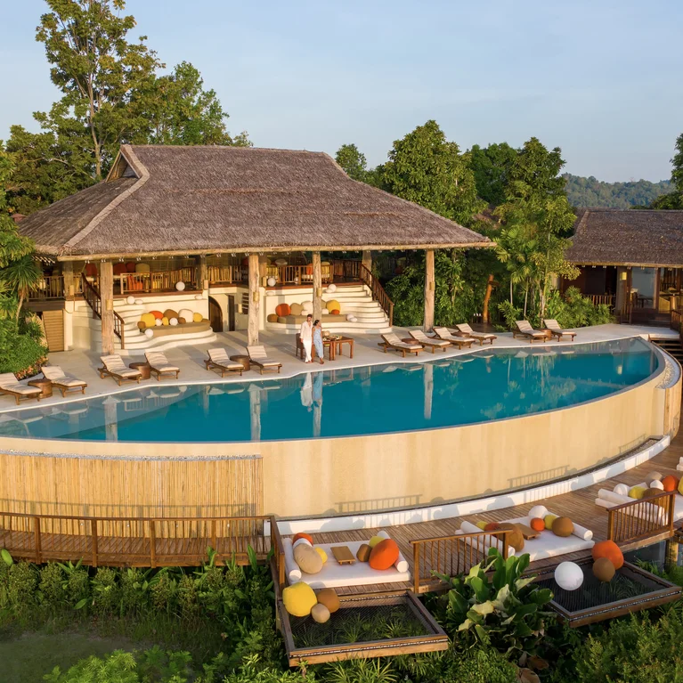 茅草屋顶别墅坐落在弧形无边际泳池旁,泳池边摆放着躺椅,开阔的天空下是原生态的绿色植物。无障碍通道将所有区域连接起来。.