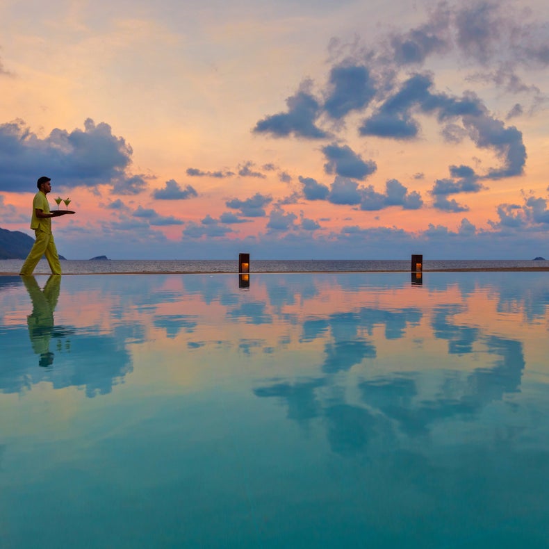 Guest enjoys tranquility beside an infinity pool at sunset, colorful sky mirrored in the water, with mountain and ocean views beyond.