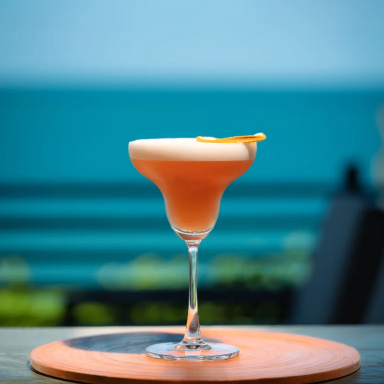 كوكتيل وردي اللون مع مقبلات قشر البرتقال موضوعة على طاولة، مع محيط هادئ وسماء صافية تشكل خلفية هادئة. الكوكتيلات البديلة متوفرة عند الطلب.