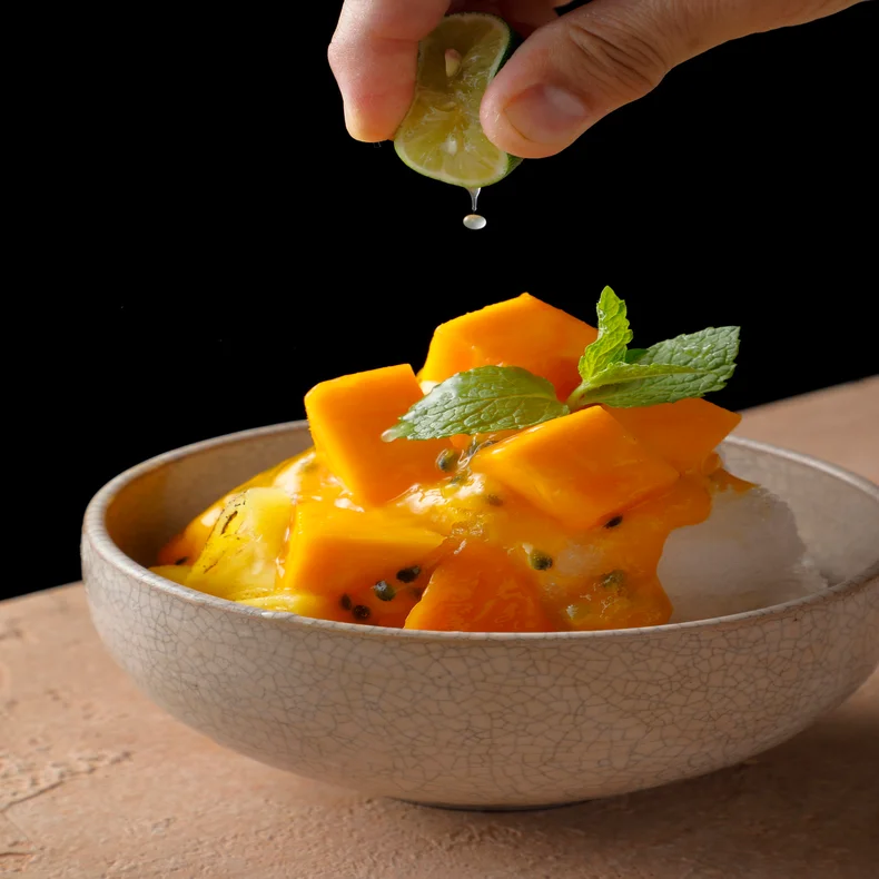 Lime juice is drizzled over shaved ice, garnished with mango pieces and mint, set against a dark background.