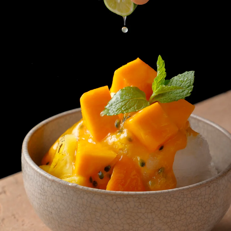 Lime juice is drizzled over shaved ice, garnished with mango pieces and mint, set against a dark background.