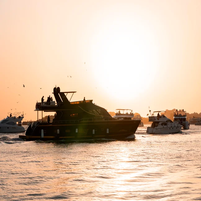 Yachts and boats glide across tranquil water at dusk, as gentle sunlight reflects softly on the surface, creating a peaceful scene.