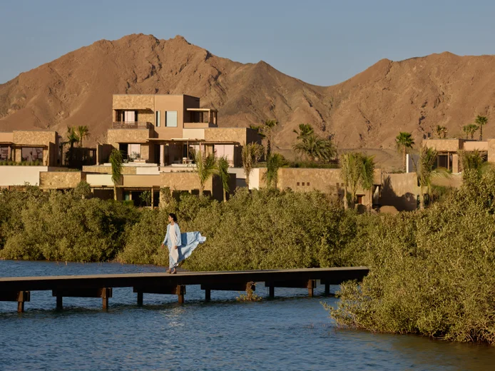 A guest in a blue dress walks along a wooden pier over water, with contemporary villas, lush greenery, and brown mountains beneath a clear sky.