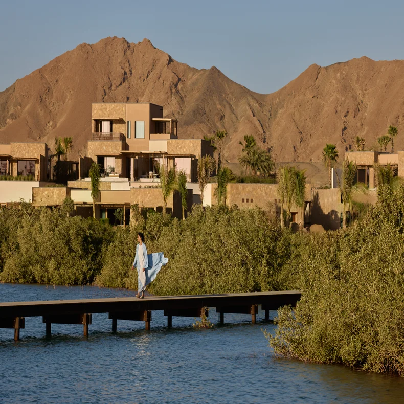 A guest in a blue dress walks along a wooden pier over water, with contemporary villas, lush greenery, and brown mountains beneath a clear sky.