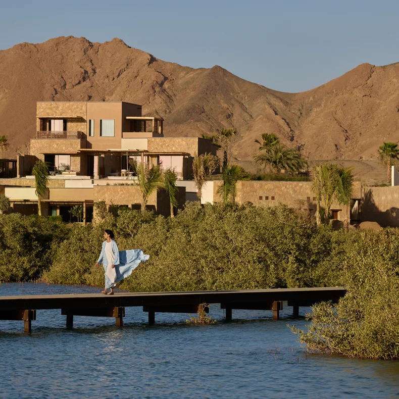 A guest in a blue dress walks along a wooden pier over water, with contemporary villas, lush greenery, and brown mountains beneath a clear sky.