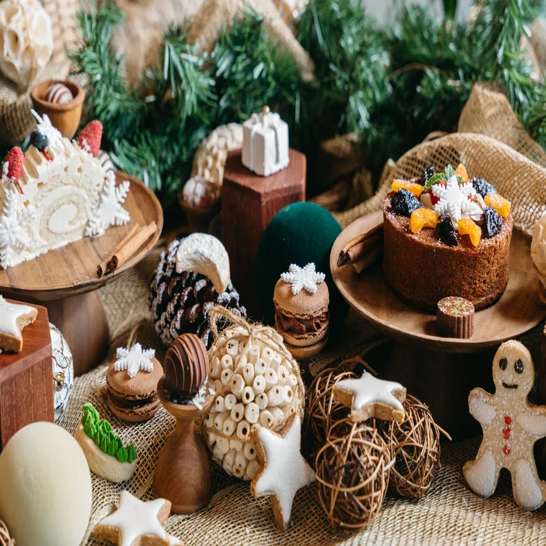 A table displays cakes, cookies, macarons, and seasonal treats on wooden stands with surrounding greenery and ornaments for a mindful holiday celebration.