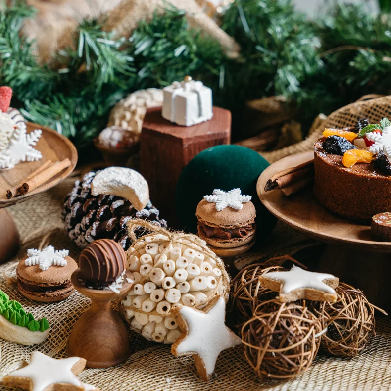 A table displays cakes, cookies, macarons, and seasonal treats on wooden stands with surrounding greenery and ornaments for a mindful holiday celebration.